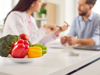 Eine Ernährungsberaterin berät einen jungen Mann. Im Vordergrund ist eine Teller gefüllt mit frischem Gemüse.