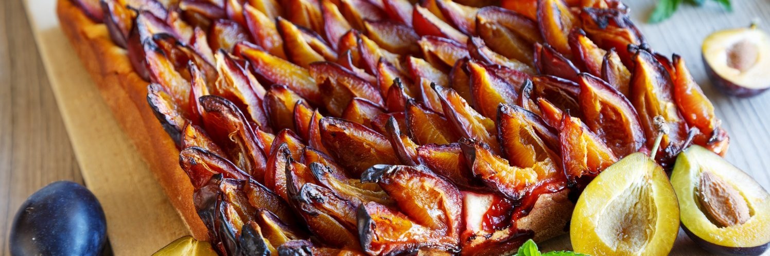 Auf einem Holzbrett liegt ein Blechkuchen mit Zwetschen, daneben liegen ganze und halbierte Früchte.