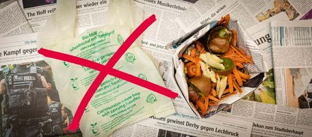 Auf verschiedenen Zeitungen liegen ein kompostierbare Muellbeutel, durchgestrichen mit einem roten Kreuz, und eine Biomuelltuete aus Zeitungspapier