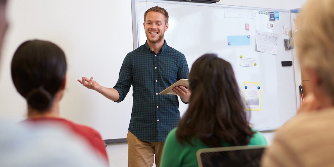 Junger Lehrer steht vor seiner Klasse, im Hintergrund ist ein Whiteboard zu sehen.. Artikel "Berufsschule" öffnen.