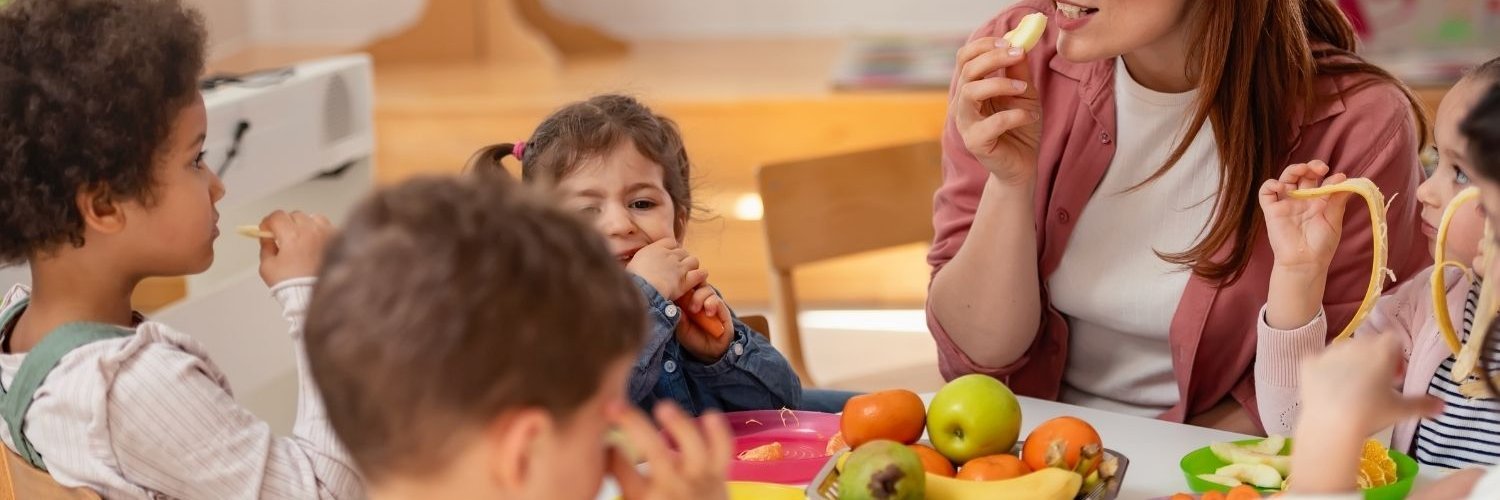 Eine Erzieherin sitzt mit drei Kita-Kindern an einem Tisch auf dem Obst und Gemüse in Stücke geschnitten steht und sie essen davon.