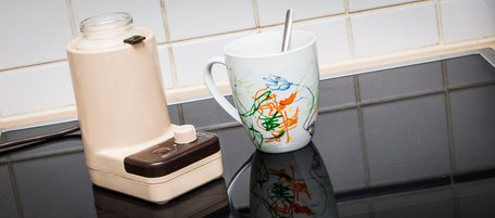 Braun-beiger Flaschenwärmer aus den 80ern wärmt ein Gläschen mit Chai-Tee, daneben eine bunt bemalte Tasse mit Löffel.