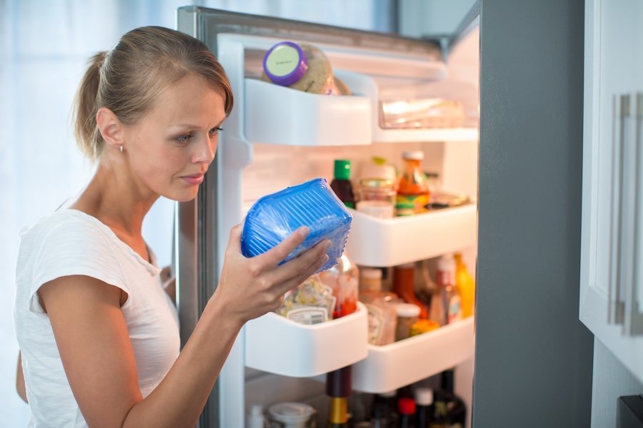 Eine Frau steht am geöffneten Kühlschrank und kontrolliert die Haltbarkeit eines verpackten Lebensmittels.