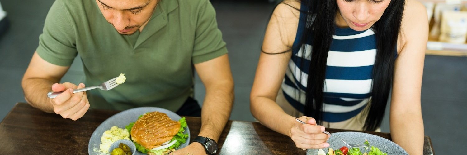 Ein Mann und eine Frau sitzen gemeinsam am Tisch und essen. Er isst einen Burger und sie einen Salat.