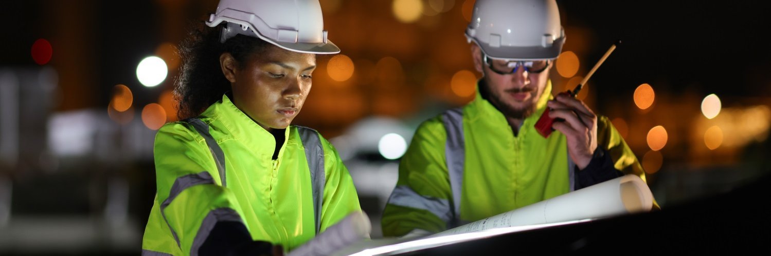 Eine Frau und ein Mann mit Sicherheitsweste und Helm beugen sich auf einer nächtlichen Baustelle über Pläne.