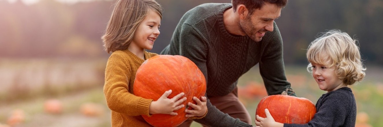 Ein Vater ist mit seinen zwei Kindern auf einem Kürbisfeld. Beide Kinder halten je einen großen Kürbis.