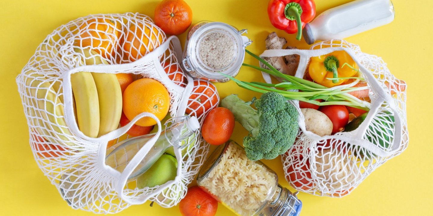 Zwei Einkausnetze auf gelben Hintergrund, gefüllt mit Obst, Gemüse und Reis, Nudeln sowie Milch in Glasgefäßen.