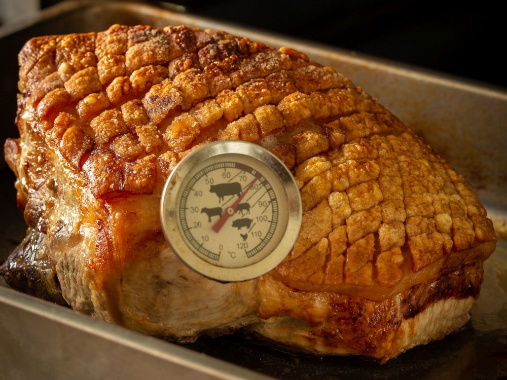 In einem knusprigen Schweinekrustenbraten steckt ein Bratenthermometer, das eine Temperatur von ungefähr 77 Grad Celsius anzeigt.