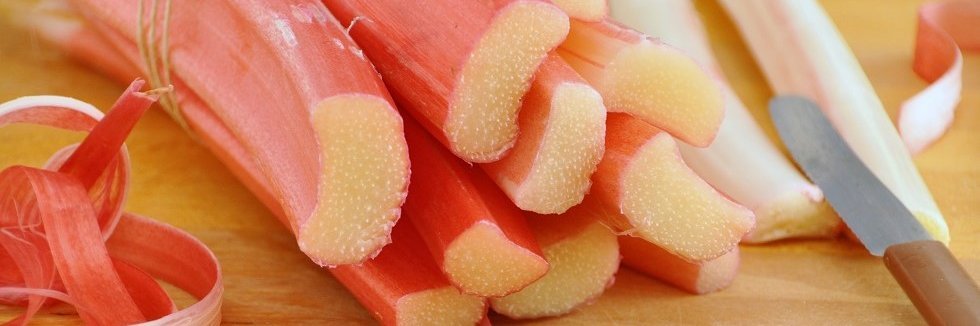 Auf einem Holztisch liegen angeschnittene Rhabarberstangen. Davor liegen Rhabarberschalen und daneben ein Messer.  