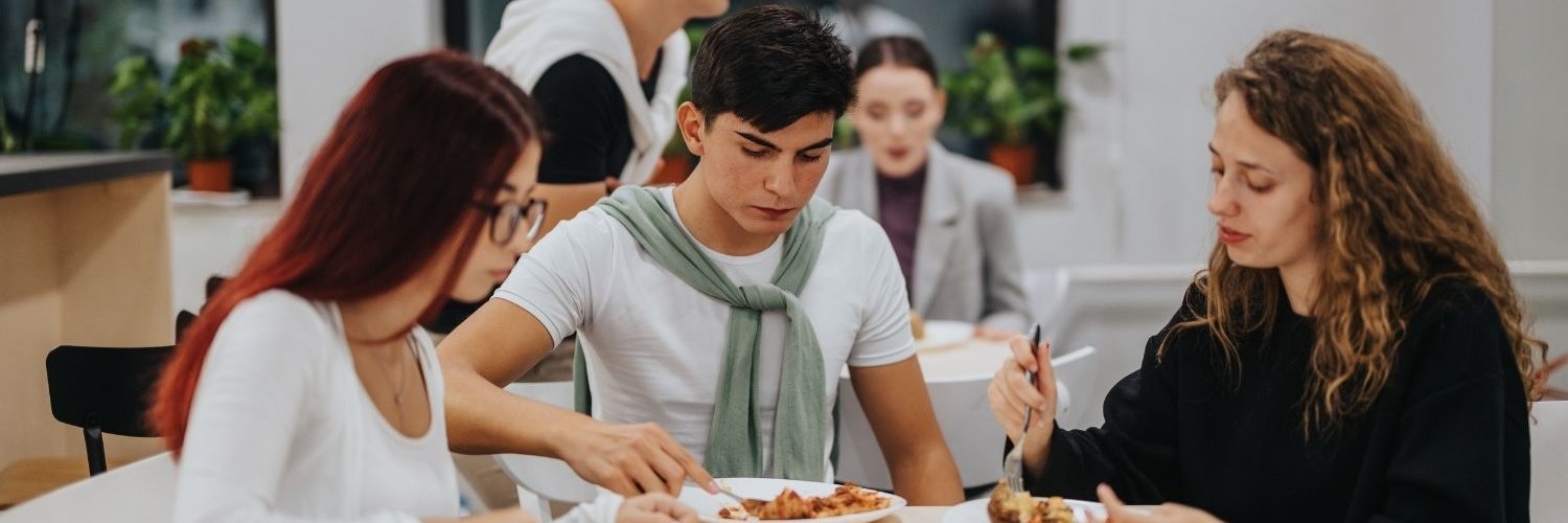 In einer Mensa sitzen zwei Studentinnen und ein Student gemeinsam beim Essen an einem Tisch.