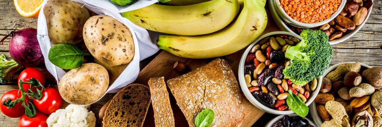 Auf einem Holztisch liegen viele ballaststoffreiche Lebensmittel wie Brot, Bananen, Kartoffeln, Linsen, Bohnen, Nüsse, Gemüse und Obst.