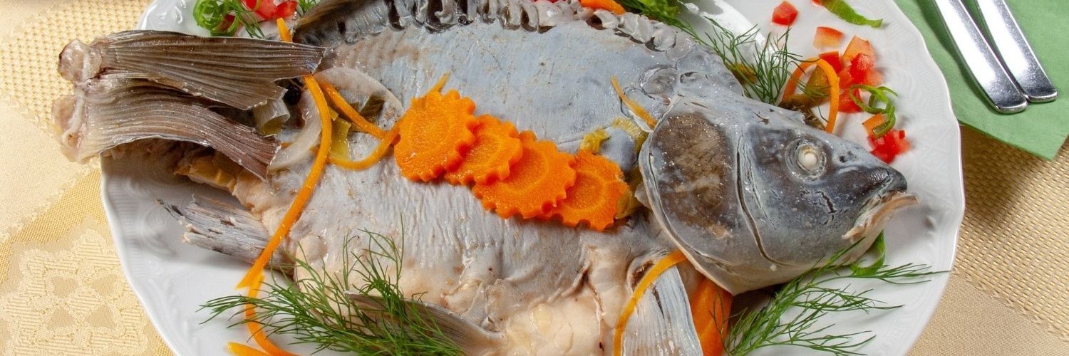 Ein ganzer gegarter Karpfen liegt auf einem Teller, garniert mit Möhren, Dill und Zitrone. 