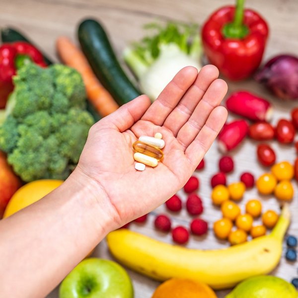 Abgebildet sind Nahrungsergänzungsmittel auf einer Hand, im Hintergrund ist Obst und Gemüse.