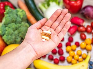 Abgebildet sind Nahrungsergänzungsmittel auf einer Hand, im Hintergrund ist Obst und Gemüse.