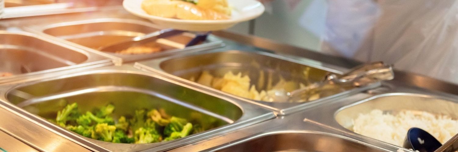 In einer Essensausgabe sind mehrere Edelstahlbehälter mit Brokkoli, Reis und Fisch gefüllt. Eine Küchenkraft steht dahinter und füllt einen Teller.