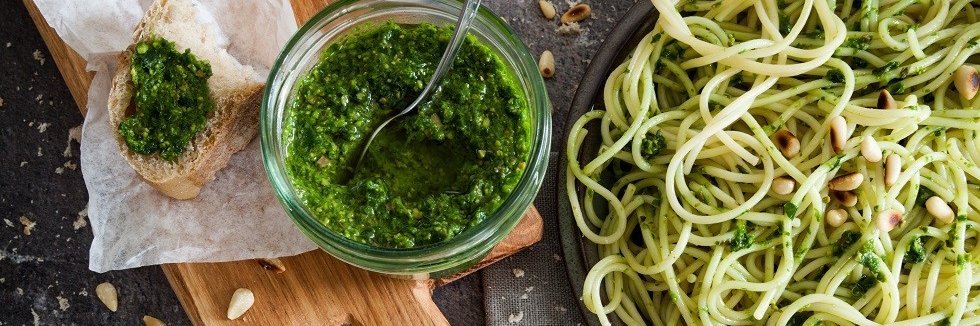 In einem Glasgefäß ist selbstgemachtes Bärlauch-Pesto. Rechts daneben steht ein Teller mit Spaghetti angerichtet mit dem Pesto. Und links daneben liegt eine Scheibe Baguette bestrichen mit dem Pesto. 