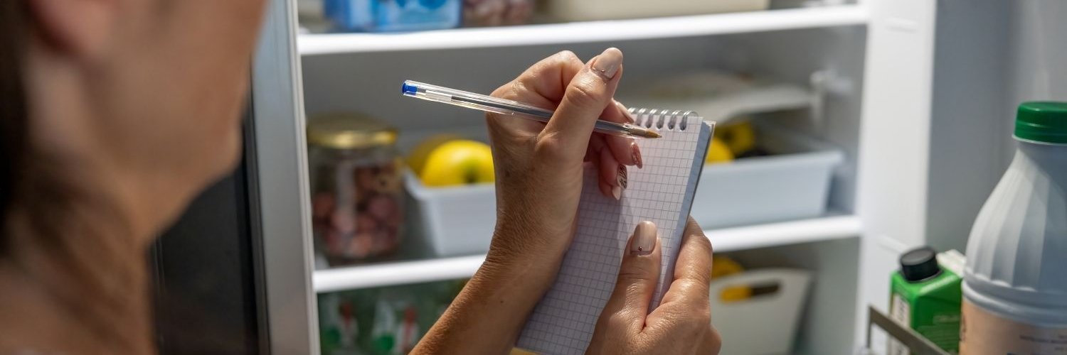 Vor einem geöffneten Kühlschrank steht eine Frau und macht sich Notizen auf einem Einkaufszettel.