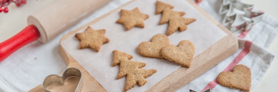 Auf einem Holzbrett liegen auf Backpapier ausgestochene Plätzchen mit Weihnachtsmotiven, daneben liegen Ausstechformen und ein Nudelholz. 