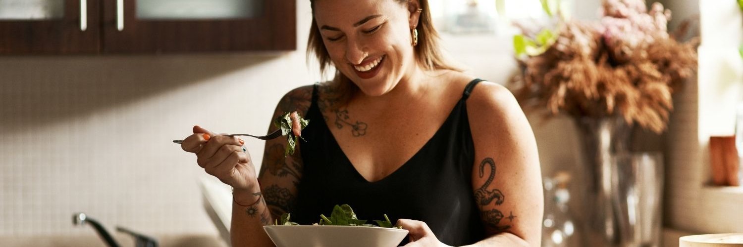 Eine junge dunkelhaarige Frau steht in einer Küche und isst Salat aus einem weißen Teller.