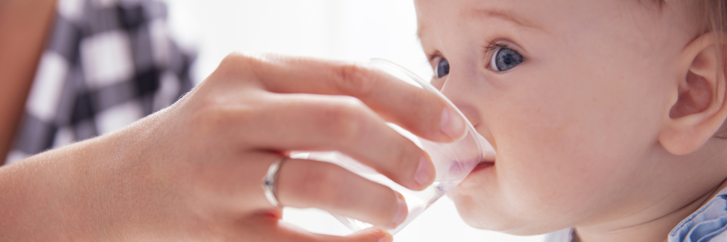 Eine Frauenhand hält einem Baby ein kleines Glas Wasser vor den Mund und es versucht zu trinken.