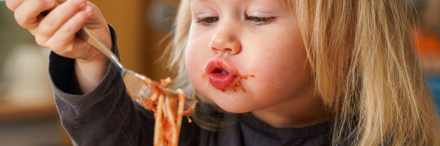 Ein blondes Mädchen im dunklen Oberteil führt mit einer Gabel etwas ungelenk Spaghetti mit Tomatensauce zum Mund und spitzt die Lippen. Ihr Mund ist mit Sauce verschmiert.