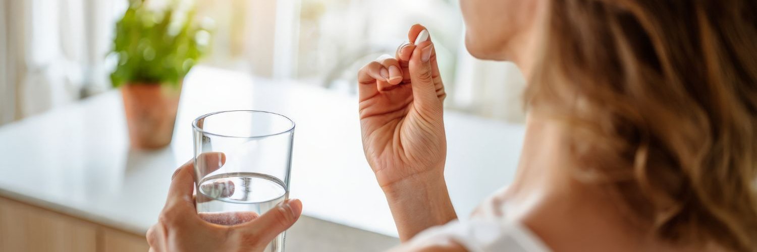 Von hinten gesehen hält eine Frau mit welligem Haar eine weiße Kapsel an ihre Lippen, während sie ein durchsichtiges Glas Wasser in der Hand hält.