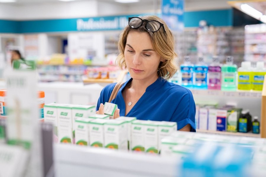 Eine Frau steht vor einem Apothekenregal und schaut sich die Verpackung eines Nahrungsergänzungsmittels an.