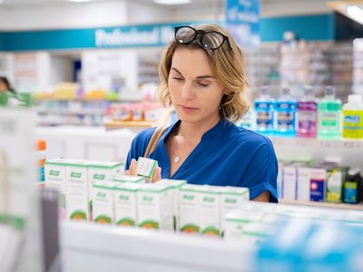 Eine Frau steht vor einem Apothekenregal und schaut sich die Verpackung eines Nahrungsergänzungsmittels an.
