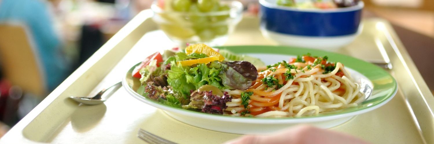 Hände tragen ein Tablett mit Essen in der Schulmensa. Auf einem Teller sind Nudeln mit Sauce und Salat, daneben seht eine Glasschale mit Trauben und eine Schale mit Gemüse. 