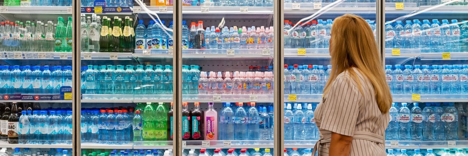 Eine Frau steht vor der großen Auswahl an Getränken im Kühlregal eines Supermarkts. 