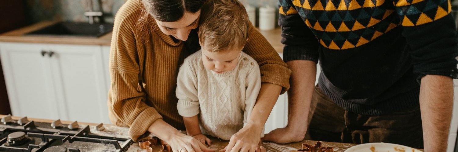 Eltern backen mit ihrem etwa zweijährigen Kind Plätzchen. Das Kind sticht mit einem Förmchen den ausgerollten Teig aus. 