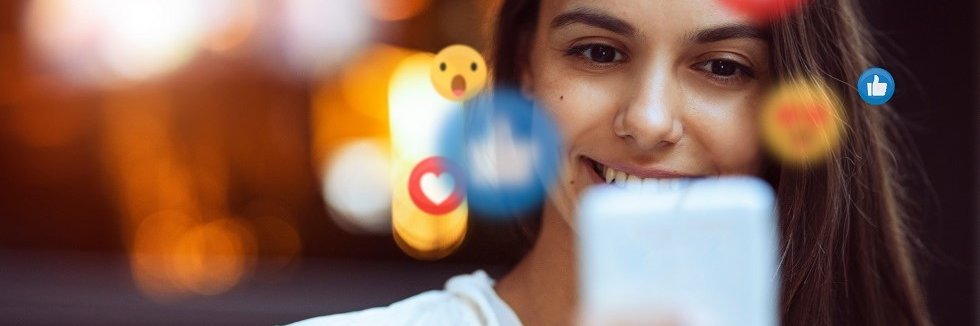 Eine junge Frau hält ein Handy in der Hand und schaut auf das Display. Um ihren Kropf schwirren Emojis von Instagram, zum Beispiel Herzen und Daumen.