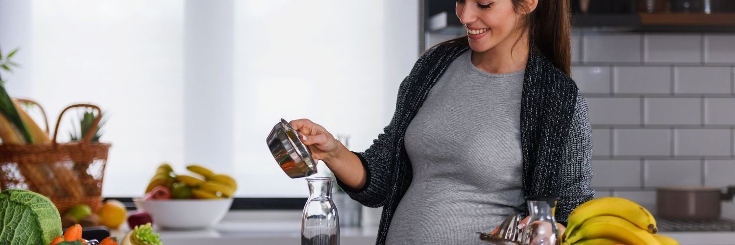 Eine junge dunkelhaarige Schwangere steht in einer Küche und gießt Orangensaft aus einer Saftpresse in eine Flasche.
