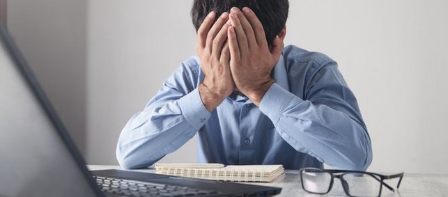 Ein Mann sitzt mit Kopfschmerzen vor seinem Laptop und hält sich seine Hände vors Gesicht