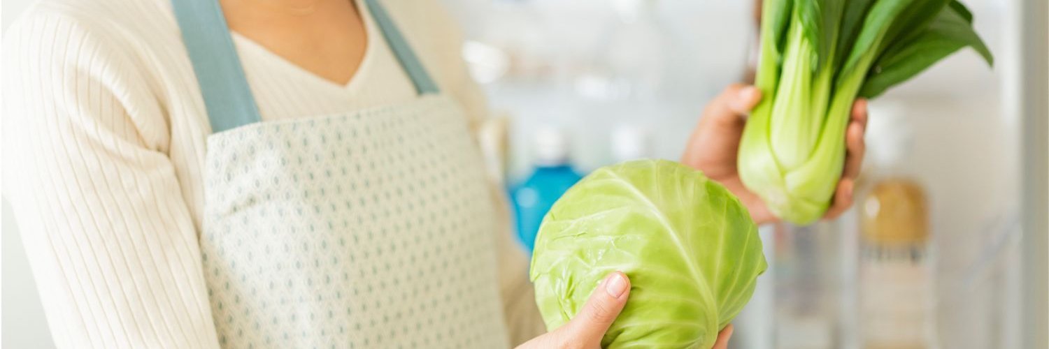 Eine Frau steht in der Küche und hält einen Fenchel und einen Weißkohl in den Händen. 