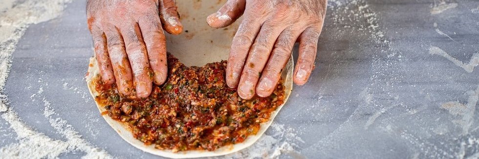 Auf einer bemehlten Arbeitsfläche liegt ein Lahmacun-Fladen. Zwei Hände verteilen darauf den Belag. 