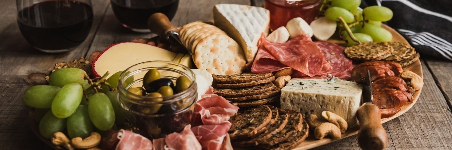 Auf einer ovalen Platte sind verschiedene Käsesorten, Schinken und Wurst mit Trauben, Nüssen, Chutney und Crackern angerichtet.
