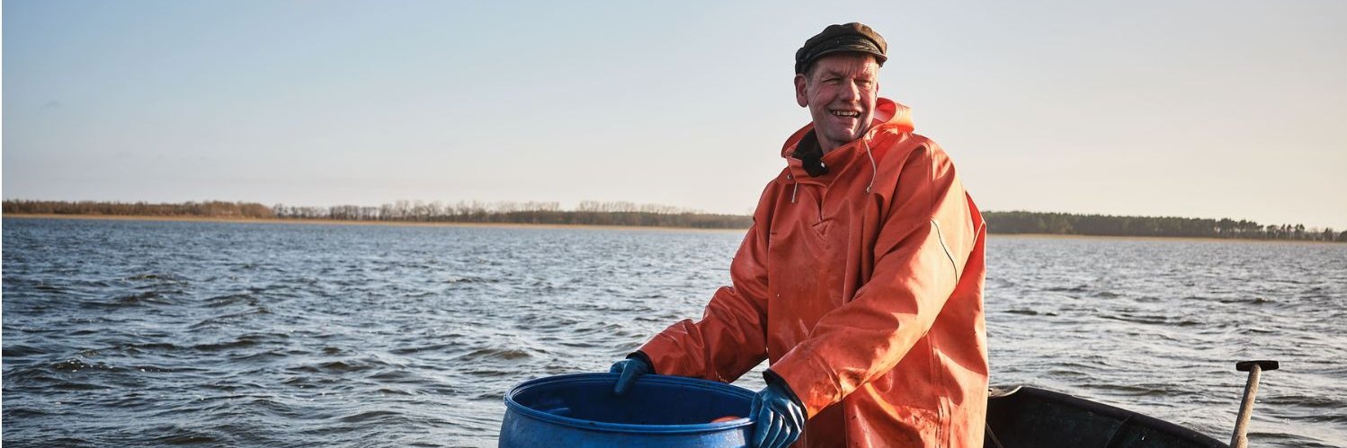 Ein älterer Mann in Watthose, oranger Regenjacke und Mütze steht in einem Ruderboot auf einem See und hält eine große blaue Tonne in seinen Händen.