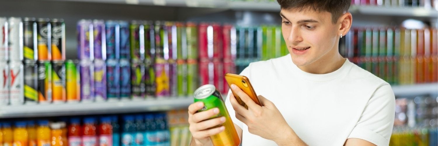 Ein junger Mann steht vor einem Getränkeregal, in der Hand eine Getränkedose und ein Smartphone, auf das er schaut.