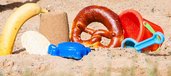 Sandkasten mit rotem Eimerchen, blauer Schaufel und Schildkröten-Förmchen sowie Banane, Brezel und Maiswaffel