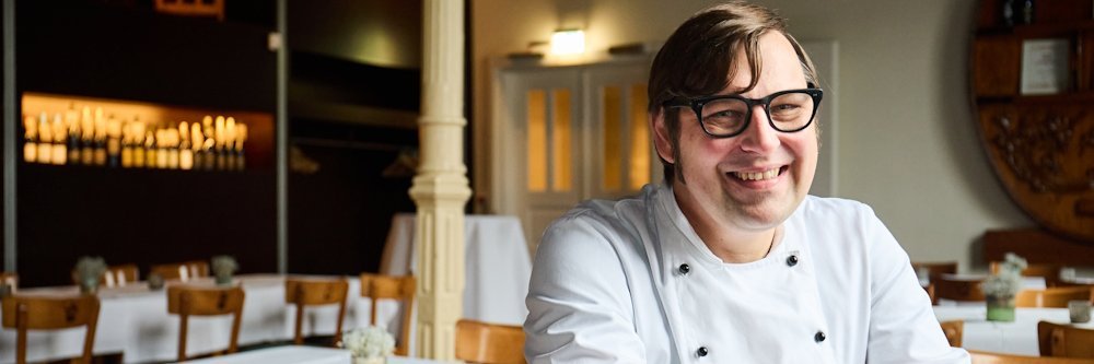 Ein Mann mit einer weissen Kochjacke sitzt rittlings auf einem Stuhl in einem Restaurant und laechelt in die Kamera.