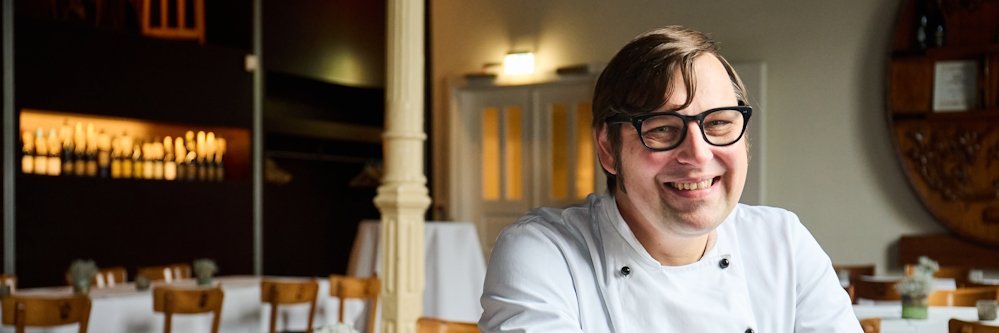 Ein Mann mit einer weissen Kochjacke sitzt rittlings auf einem Stuhl in einem Restaurant und laechelt in die Kamera.