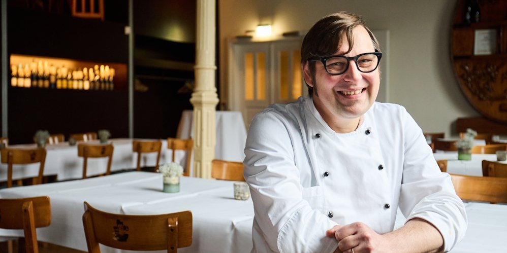Ein Mann mit einer weissen Kochjacke sitzt rittlings auf einem Stuhl in einem Restaurant und laechelt in die Kamera.. Artikel "Im Bio-Restaurant „Zur Erholung"" öffnen.