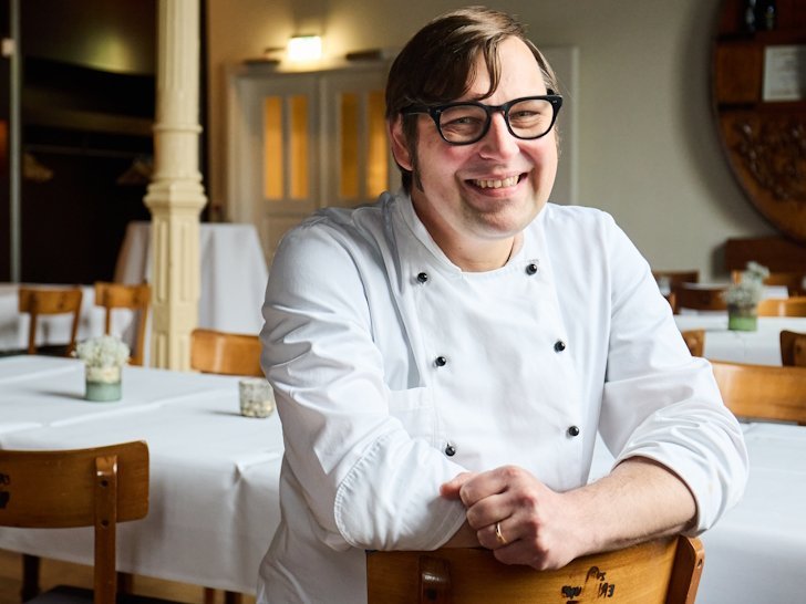 Ein Mann mit einer weissen Kochjacke sitzt rittlings auf einem Stuhl in einem Restaurant und laechelt in die Kamera.