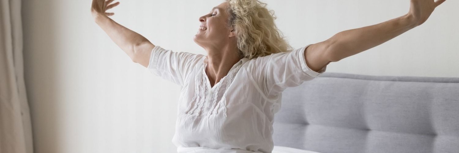 Eine ältere Frau sitzt im Bett und streckt genüsslich die Arme zu beiden Seiten aus.