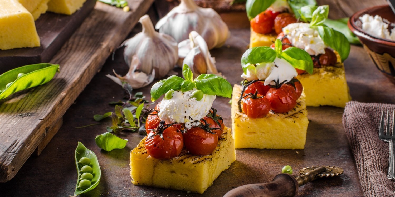 Auf einem dunklen Untergrund liegen gegrillte Polentastücke mit gebackenen Kirschtomaten, Mozzarella, Basilikum und Erbsen angerichtet.  . Artikel "Polenta – das Maisgericht aus Italien" öffnen.
