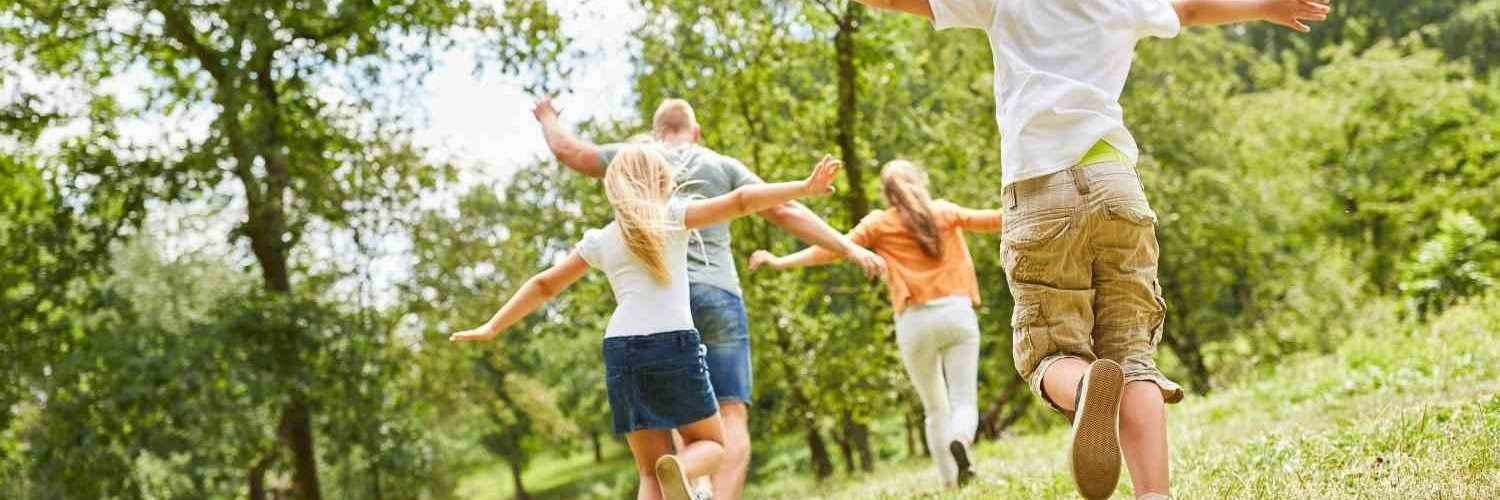 Auf einer Waldlichtung laufen ein Junge und ein Mädchen mit ausgebreiteten Armen hinter ihren Eltern her.