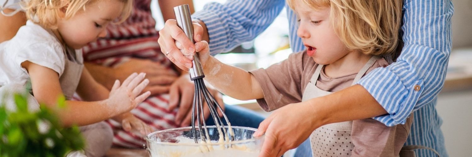 In einer Küche führt eine Mutter die Hand eines blonden Kleinkindes beim Rühren von Teig. Ein anderes Kind sitzt mit bemehlten Händen auf der Arbeitsfläche beim Vater.