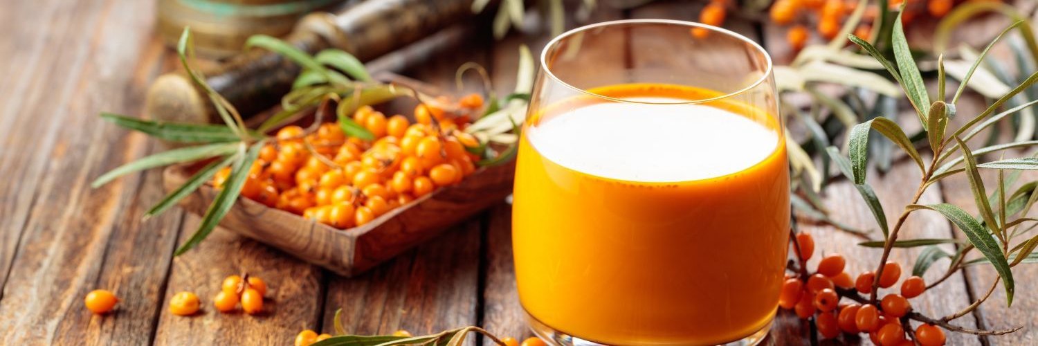 Auf einem groben Holztisch steht ein Glas mit leuchtend orangenem Sanddornsaft. Daneben liegen Sanddornbeeren und Zweige.