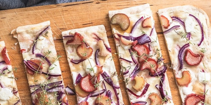 Flammkuchen mit Rhabarber, roten Zwiebeln, Ziegenkäse und Honig liegt in Stücke geschnitten auf einem Holzbrett. . Artikel "Rhabarber herzhaft genießen" öffnen.